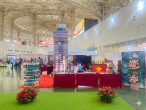 Hình ảnh gian hàng lớn, hiện đại của Công ty Cổ phần Đầu tư và Thương mại Quốc tế VCC tại triển lãm Vietbuild 2026. Banner chính ghi 'VCC: CHẤT LƯỢNG - UY TÍN - BỀN VỮNG' và thông tin liên hệ. Các màn hình lớn trình diễn sản phẩm. Rất nhiều khách tham quan và nhân viên tư vấn. Foreground có cỏ nhân tạo và chậu cây hoa hồng trắng.