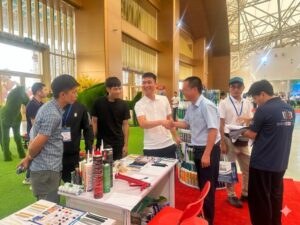 Nhân viên VCC mặc áo sơ mi xanh đang bắt tay thân thiện với khách hàng tại gian hàng triển lãm Vietbuild 2026. Trên bàn trưng bày có súng bắn keo màu đỏ, các tuýp keo silicone VCC và bảng mẫu sản phẩm. Một nhóm khách hàng đang tập trung tìm hiểu và thảo luận về các giải pháp chất kết dính trong không gian gian hàng hiện đại có thảm cỏ xanh nhân tạo.