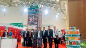 Một nhóm lãnh đạo Hội Vật liệu xây dựng (VLXD) mặc comple đứng chụp ảnh lưu niệm trước gian hàng triển lãm của Công ty VCC tại triển lãm Vietbuild 2026. Gian hàng nổi bật với tháp trưng bày cao "VCC Vì Chất Lượng Cuộc Sống", quầy trưng bày keo silicone hình tròn nhiều tầng và banner "Vật Liệu Xây Dựng Xanh". Cấu trúc mái giàn không gian kim loại lớn phía trên bối cảnh.