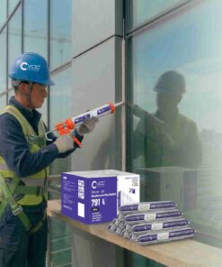 Một công nhân xây dựng đeo mũ bảo hộ màu xanh và dây an toàn đang sử dụng súng bắn keo thi công vách kính tòa nhà cao tầng với sản phẩm Keo Silicone VCC 791. Hình ảnh cho thấy hộp các-tông và các tuýp keo VCC 791 Silicone Weatherproofing, cùng với văn bản quảng cáo nêu rõ 5 ưu điểm: Chống thời tiết vượt trội, độ bền 20 năm, độ bám dính cao, kháng nước, và an toàn môi trường.