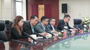 Five professionals seated at a long conference table during a formal meeting, each with a microphone in front of them and beverages nearby, in a corporate boardroom with a floral arrangement at the far end.