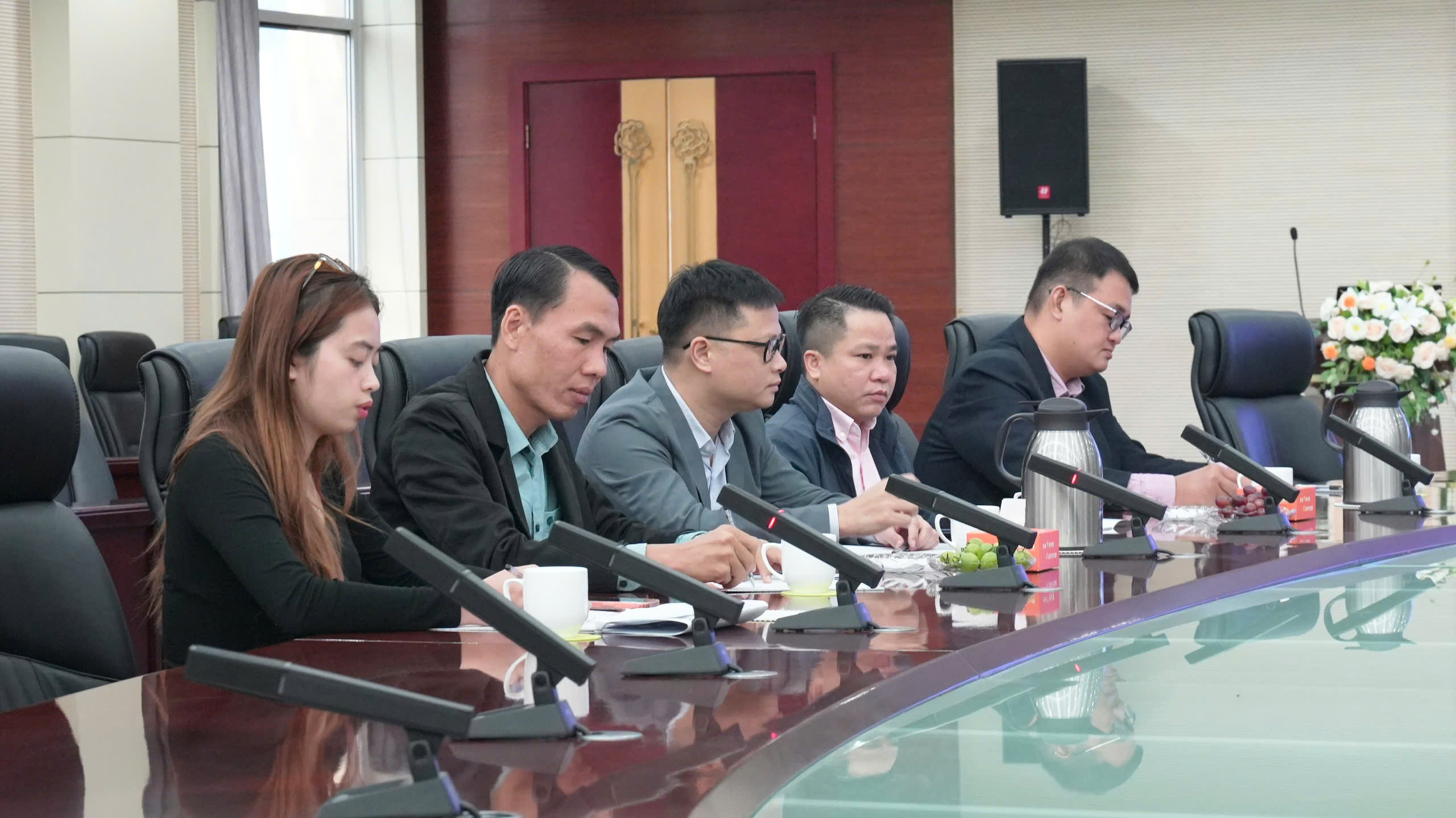 Five professionals seated at a long conference table during a formal meeting, each with a microphone in front of them and beverages nearby, in a corporate boardroom with a floral arrangement at the far end.