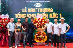 Group of Men Posing with a Large Flower Bouquet on a Red Carpet at a Grand Opening Backdrop with Vietnamese Text  Vcc Sealant   Sản Xuất Phân Phối Keo Silicone Và Chất Kết Dính Chuyên Nghiệp