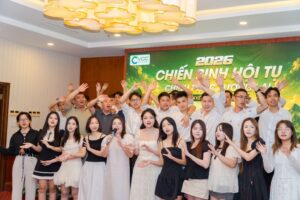 Group of young adults posing for a group photo at a 2026 event, front-row women in dresses, back-row men in white shirts, waving hands, with a green banner reading 2026 in the background.
