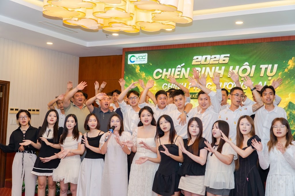 Group of young adults posing for a group photo at a 2026 event, front-row women in dresses, back-row men in white shirts, waving hands, with a green banner reading 2026 in the background.
