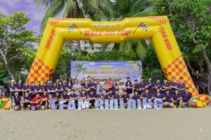 Group of People in Purple Shirts Posing on a Beach During a Team building Event Under a Yellow Inflatable Arch That Says race on Top with team Building Letters in Front  Vcc Sealant   Sản Xuất Phân Phối Keo Silicone Và Chất Kết Dính Chuyên Nghiệp