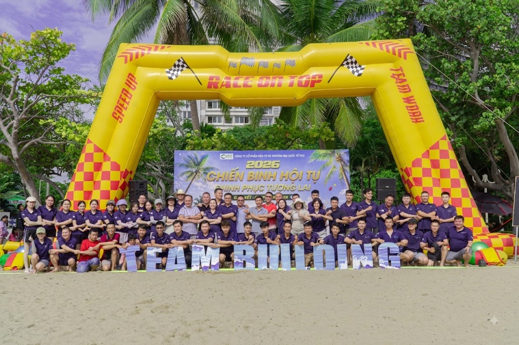 Tập thể VCC chụp ảnh kỷ niệm tại hoạt động team building VCC bãi biển 2026 – VCC Sealant - Sản xuất, phân phối keo silicone và chất kết dính chuyên nghiệp Group of People in Purple Shirts Posing on a Beach During a Team building Event Under a Yellow Inflatable Arch That Says Vcc Sealant Sản Xuất Phân Phối Keo Silicone Và Chất Kết Dính Chuyên Nghiệp'Race on Top' with 'TEAM BUILDING' letters in front.