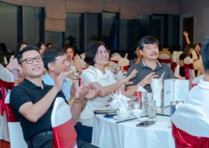 People seated at a banquet table clap and smile during a formal dinner event, white table settings and red chair covers visible in the background.