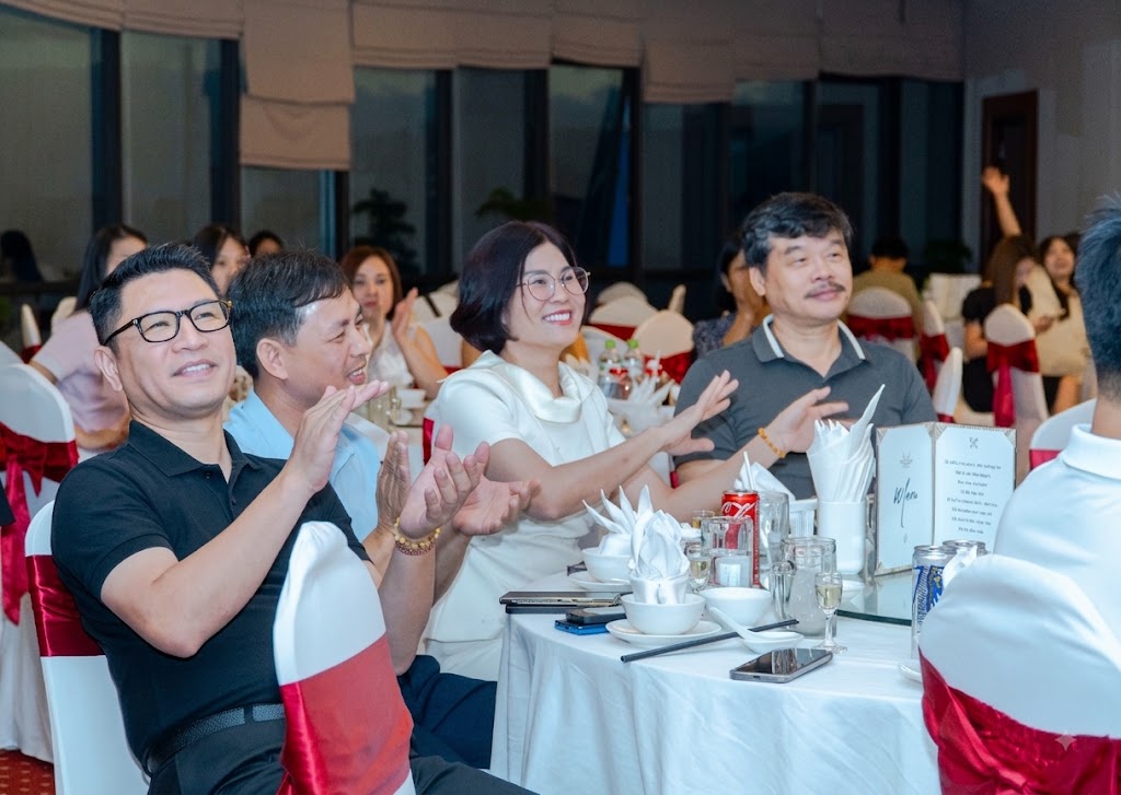 People seated at a banquet table clap and smile during a formal dinner event, white table settings and red chair covers visible in the background.