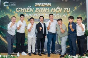 Nine men stand in a row, fists raised, posing in front of a banner that reads 2026 and Vietnamese event text, at a corporate gathering.
