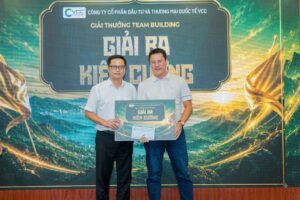 Two men in light shirts pose on stage, holding a framed certificate during an awards ceremony with a Vietnamese backdrop behind them.