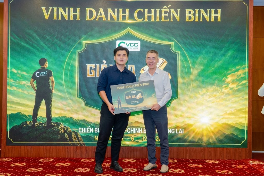 Two men on a red-carpet stage hold a certificate in front of a large green awards backdrop reading 'VINH DANH CHIẾN BINH'.